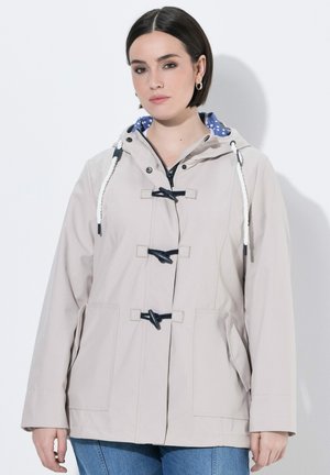 Femme portant une veste beige à capuche avec des fermetures à boutons à bascule et une doublure à pois bleus, associée à un jean bleu, debout devant un fond blanc.