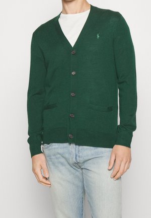 Mann trägt eine dunkelgrüne, geknöpfte Strickjacke über einem weißen Hemd und hellblauen Jeans, steht vor einem schlichten Hintergrund.