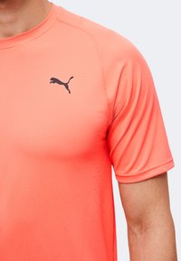 Κοντινό πλάνο ενός προσώπου που φοράει ένα φωτεινό κοραλί αθλητικό μπλουζάκι Puma με κοντά μανίκια και μικρό μαύρο λογότυπο Puma στο στήθος.