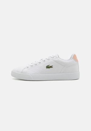 Zapatilla de lona blanca con punta redonda, cierre con cordones y un logo bordado en verde. Presenta una textura suave y un acento en el talón de color rosa claro.