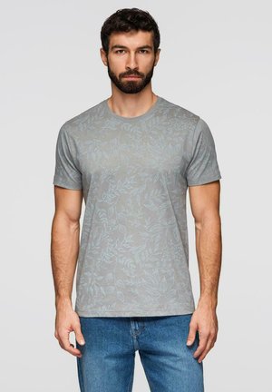 Mann mit dunklen Haaren und Bart, trägt ein graues kurzärmeliges T-Shirt mit einem dezenten hellblauen Blättermuster und blaue Jeans, steht und schaut nach vorne.