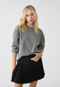 Grau gestrickter Pullover mit geripptem Kragen und umgeschlagenen Bündchen, kombiniert mit einem schwarzen Faltenrock. Fokus auf Textur und Schichtung der Kleidungsstücke.