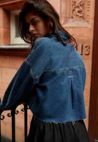 Giacca di jeans blu scuro, dal design accorciato con orlo sfrangiato, cuciture dettagliate e colletto classico. Indossata su una gonna nera a ruota.