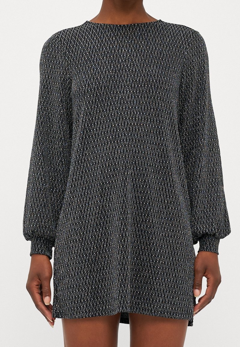 Femme portant une mini-robe noire à manches longues avec des motifs zigzag argentés et des poignets froncés, debout devant un fond uni.