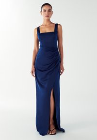 Moteris vilkinti be rankovių tamsiai mėlynos satino suknelę su kvadratine iškirpte ir priekinėmis iškirpėmis, stovi prieš paprastą baltą foną.