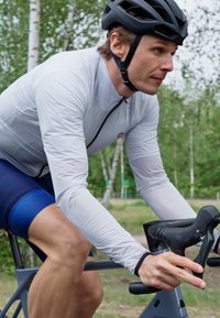 Cycliste portant un maillot à manches longues gris clair et un short marine, casqué d'un casque noir, tenant des poignées noires sur un vélo dans un cadre naturel.