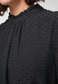 Black blouse featuring a high neckline with subtle ruffle trim, textured fabric with raised dot patterns, and pleats along the front.