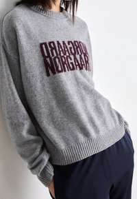 Maglione grigio a maglia con orlo e polsini a coste, caratterizzato da audaci grafiche testuali bordeaux sulla parte anteriore. Tessuto morbido, vestibilità rilassata.