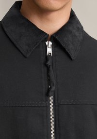 Schwarze Jacke mit einem Cordkragen, Frontreißverschluss und strukturiertem Stoff. Das Design umfasst eine glatte, strukturierte Passform und minimale Nähdetails.