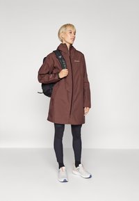 Brun vanntett jakke med høy krage, hel glidelås og sidelommer. Brukt over svarte leggings og kombinert med joggesko.