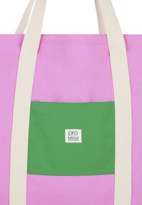 Roze canvas draagtas met een groene patchzak. De tas heeft crèmekleurige banden en een klein wit label met de tekst "pro test".