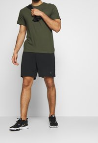 Camiseta verde oliva de manga corta, pantalones cortos negros con el logo de Nike, zapatillas negras con suelas blancas, de pie sobre un fondo sencillo.