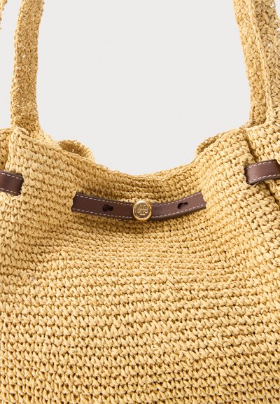 Μπεζ υφασμάτινη τσάντα από άχυρο με δύο λαβές και καφέ δερμάτινο λουρί που διαθέτει ένα μικρό στρογγυλό χρυσό κουμπί.