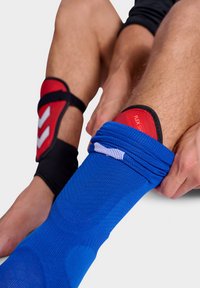 Blå sportstrumpa med textilstruktur, utrustad med ett svart ankelstöd och röd undersida märkt "FLEX SHIELD." Nära bild av fot i rörelse.