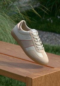 Baskets beiges et blanches avec des rayures marron clair reposant sur un banc en bois à l'extérieur, avec un arrière-plan d'herbe et de gravier.