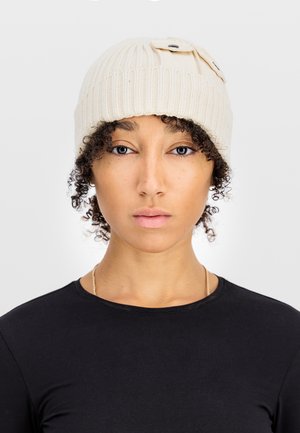Woman with curly hair wears a cream knit beanie with buttons on top and a black crew-neck shirt against a plain white background.