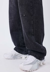 Pantaloni in denim neri con una vestibilità rilassata, cuciture dettagliate e tasche laterali; abbinati a sneakers bianche Nike con suola ammortizzata.