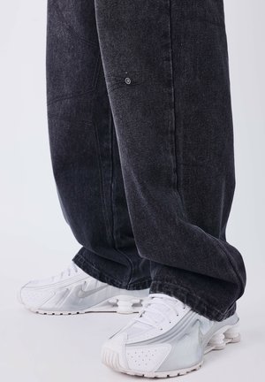 Pantalones de mezclilla negros con un ajuste relajado, costuras detalladas y bolsillos laterales; combinados con zapatillas Nike blancas que cuentan con una plantilla acolchada.