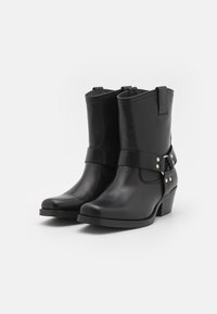 Botas ankle de cuero negro con puntera redondeada, correa decorativa y acentos en metal plateado, con un tacón bajo en bloque y costura frontal.