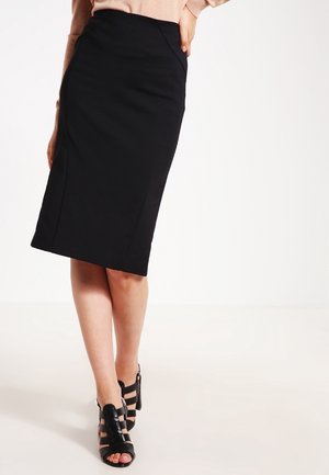 Woman wearing a black knee-length pencil skirt and black strappy high-heeled sandals, standing against a plain white background.