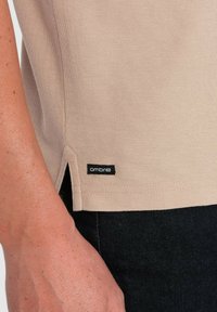 Béžový pletený top s texturovanou tkaninou, bočními rozparky a černou „ombre“ etiketou, kombinovaný s tmavými džínami. Zaměření na materiál a designové detaily.