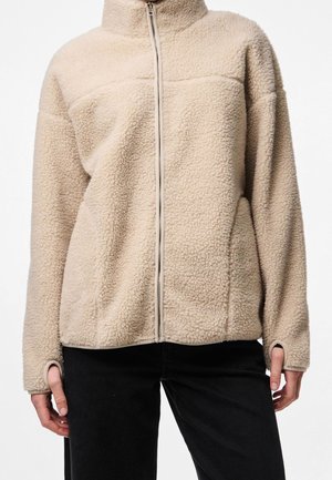 Person iført beige fleecejakke med lynlås og hul til tommelfingrene, kombineret med sorte bukser, vist fra skuldre til hofter.