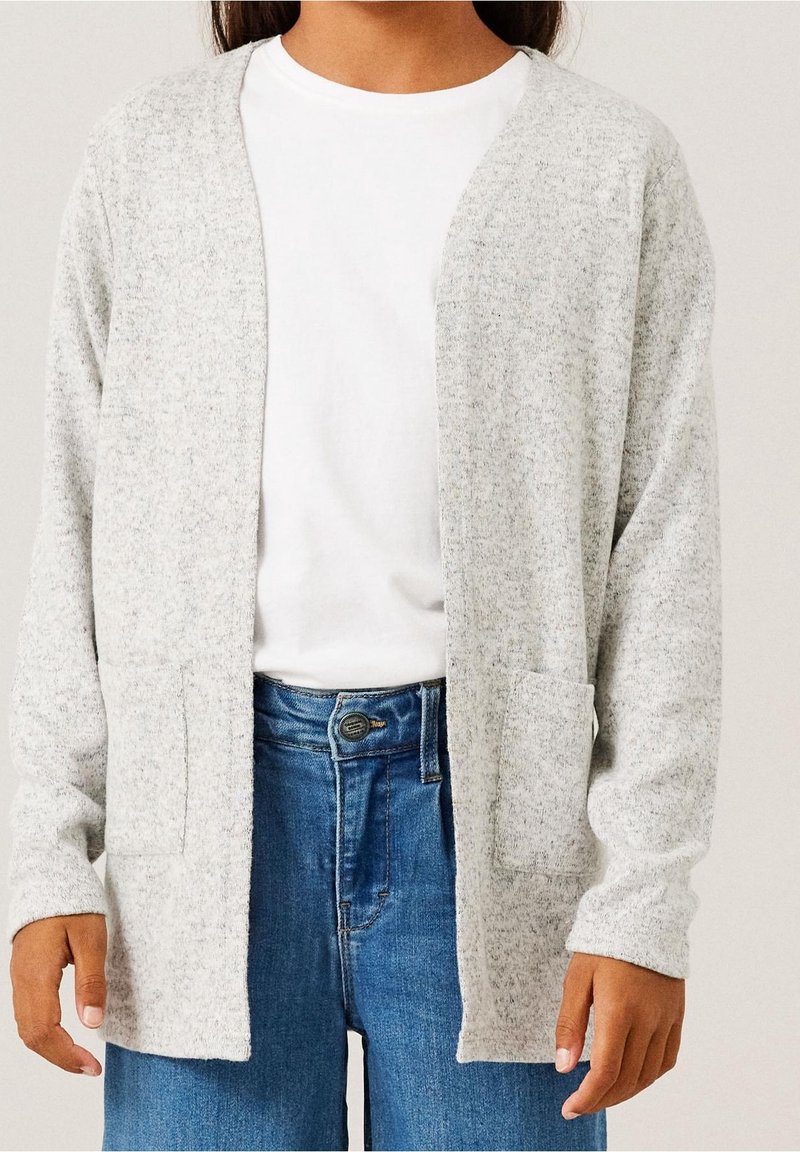 Personne portant un cardigan gris clair ouvert sur un T-shirt blanc avec un jean bleu, mains relâchées le long du corps, fond neutre.