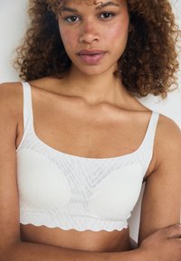 Ung kvinna med lockigt hår som bär en vit mönstrad bralette, står mot en enfärgad ljus bakgrund med armarna i kors.