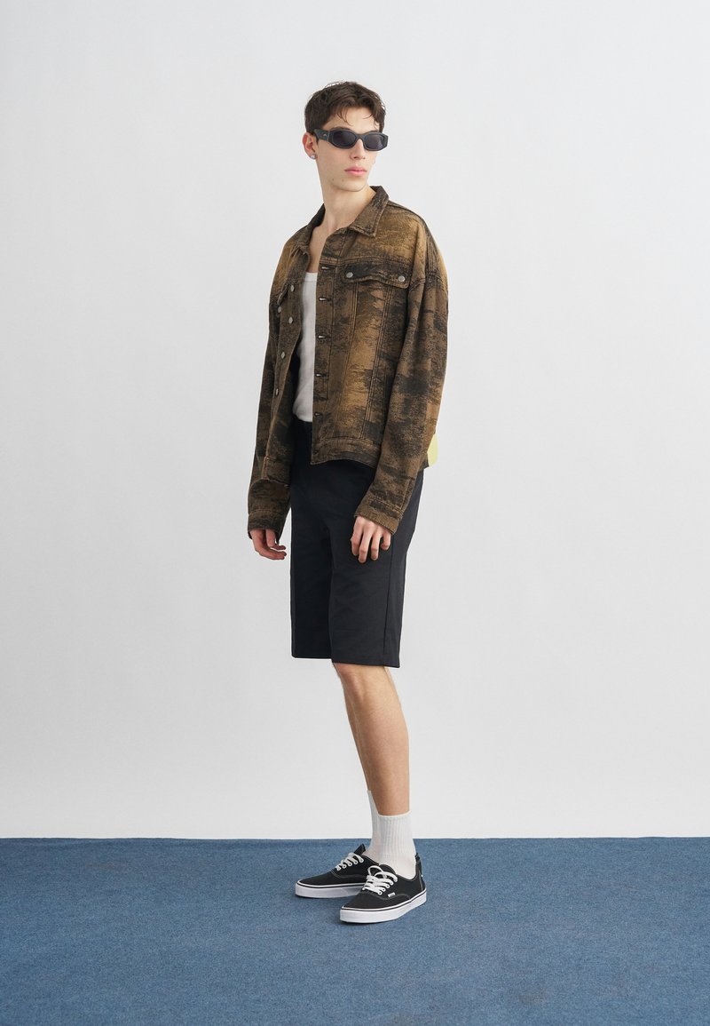 Giovane uomo con occhiali da sole neri, giacca marrone con motivo, canottiera bianca, pantaloncini neri, calzini bianchi e scarpe da ginnastica nere in piedi su un tappeto blu.