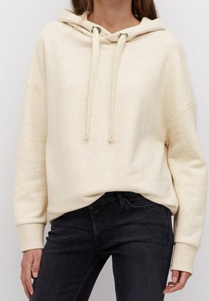 Person iført en cremefarvet hættetrøje med snore og mørke jeans med hænderne afslappet ved siden.