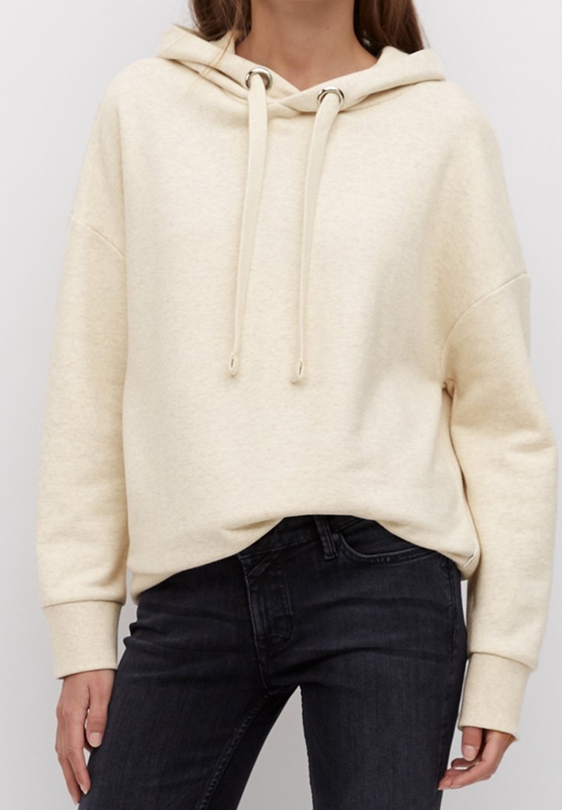 Personne portant un sweat à capuche couleur crème avec des cordons et un jean foncé, les mains relâchées sur les côtés.