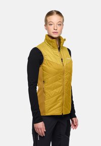 Gelbe isolierte Weste mit hohem Kragen, mit Zickzacksteppung und seitlichen Taschen, kombiniert mit einem schwarzen Langarmshirt.