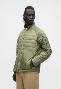 Muž v svetlo zelenej prešívanej bunde s náprsným vreckom na rukáve a béžových nohaviciach, pozerá sa doľava na bielom pozadí.