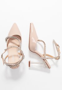 Nude patentläderklackar med spetsig tå, juvelbesatta ankelremmar och genomskinliga blockklackar, med en slät textur och en elegant design.