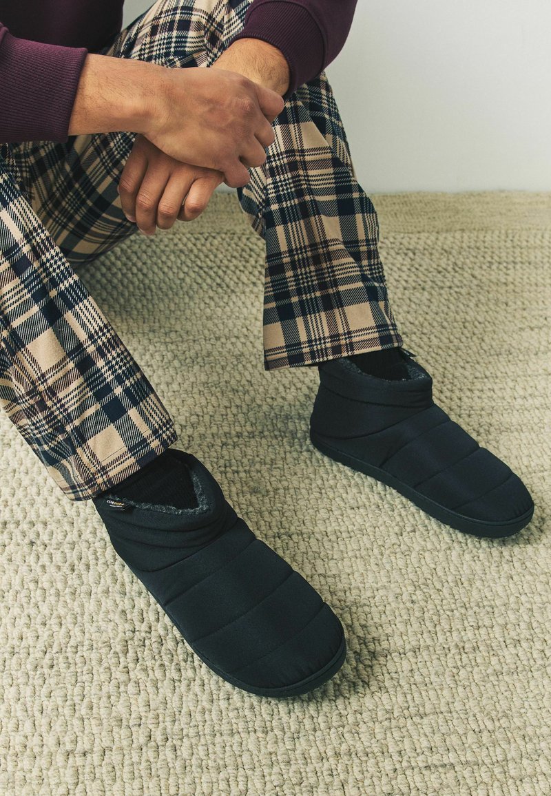 Juodos siuvinėjimo šlepetės su švelniu pūkuotu pamušalu ir guminiais padais, derinamos su smėlio ir tamsiai mėlynomis languotomis kelnėmis ant tekstūruoto pilko kilimo.