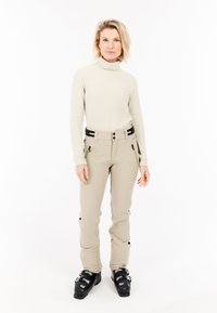 Vrouw in beige skibroek, crème coltrui en zwarte skischoenen staande tegen een witte achtergrond.