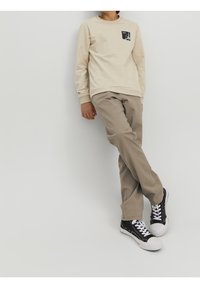 Beige långärmad skjorta med en svart grafisk ficka, tillsammans med ljusbruna byxor och svarta sneakers med vita snören och tåhättor.