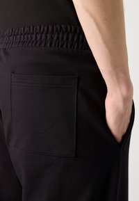 Black elastic-waist pants feature a flat pocket on the back. The fabric is smooth with a soft texture, highlighting a simple design.