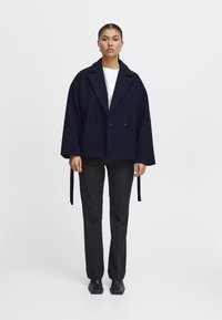 Giacca doppiopetto blu navy con maniche ampie e lacci laterali, abbinata a pantaloni neri a gamba dritta e stivaletti alla caviglia con punta quadrata.