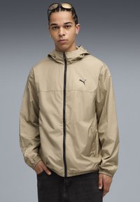 Lyst beige hættejakke med en sort lynlås, elastiske manchetter og sidelommer. Har et lille sort logo på brystet.
