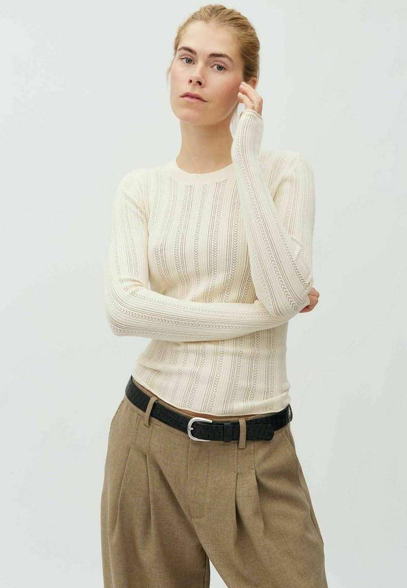 Femme aux cheveux blonds portant un pull en maille crème à manches longues et un pantalon plissé marron taille haute avec une ceinture noire.