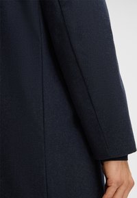 Dettaglio di un cappotto blu navy con tessuto texturizzato, manica strutturata e cuciture pulite, caratterizzato da una finitura leggermente lucida. Progettato per un look sartoriale.