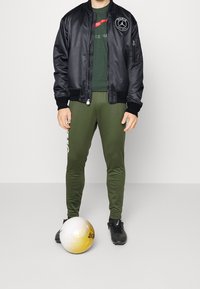 Black satin bomber jacket with logo, green t-shirt, green fitted pants, and black athletic shoes, standing next to a white and gold soccer ball.