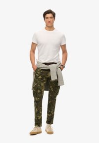 Witte crewneck t-shirt, olijfgroene camouflage cargobroek met meerdere zakken, lichtgrijze trui om de taille geknoopt, beige sneakers.