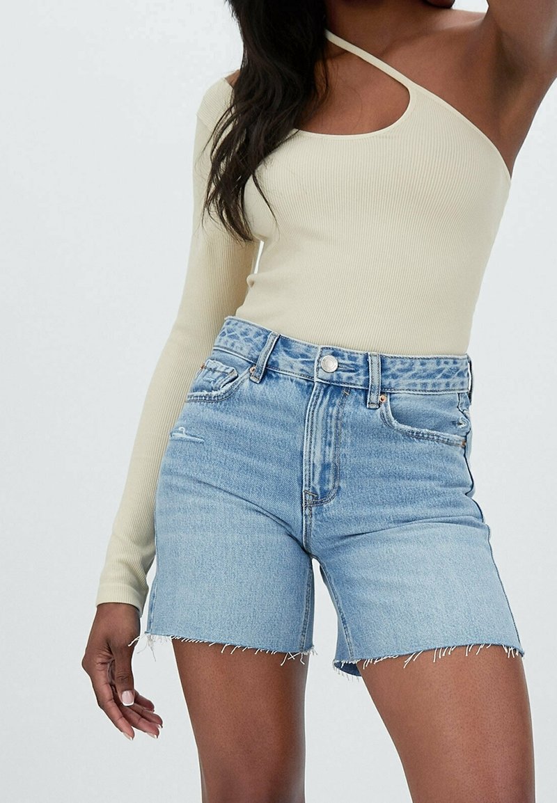 Femme portant un short en denim bleu clair avec des ourlets effilochés et un top crème à manches longues et à une épaule sur un fond uni.