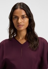 Donna con capelli scuri alla spalla che indossa una maglietta scollata a V di un viola intenso, guarda dritto in avanti con un'espressione neutra.