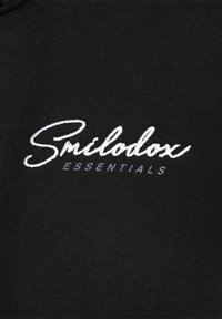 Tissu noir avec une texture lisse comportant la broderie "Smilodoux ESSENTIALS" en blanc. Le texte présente une combinaison de lettres cursives et de lettres en bloc.