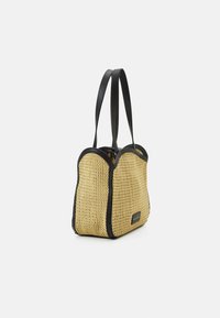 Sac fourre-tout rectangulaire en paille tressée avec une bordure noire et deux longues bretelles noires, présenté légèrement en angle sur fond blanc.