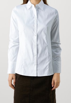 Woman wearing a fitted white long-sleeve button-up shirt and a dark brown skirt, standing against a plain background.