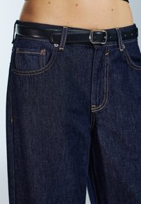 Tamnoplave traperice ravnog kroja s kontrastnim šavovima i crnim kožnim remenom sa srebrnom kopčom. Prikazani su i prednji džepovi.